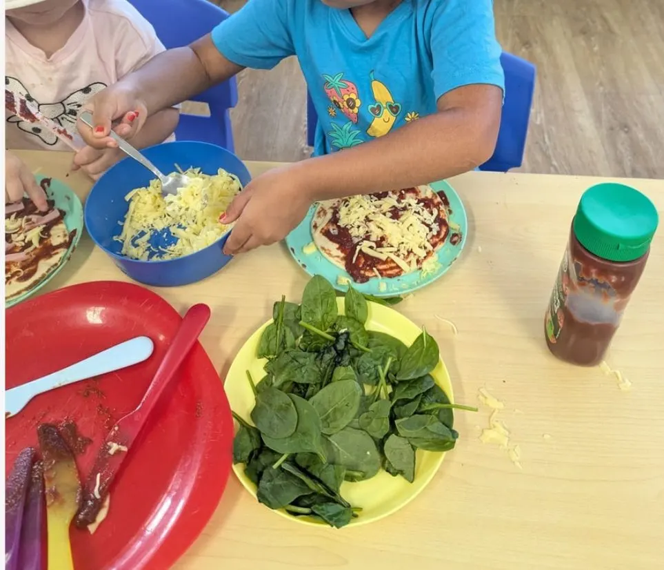 Children topping their own pizzas with cheese, sauce and fresh spinach at the table