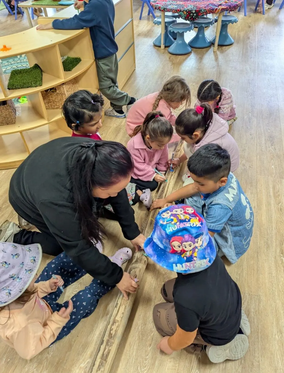 An educator and a group of children drawing together on a long timber log indoors